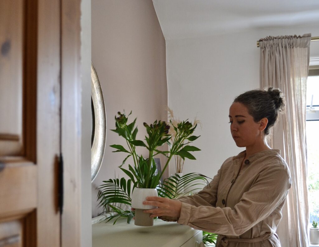 Zoe is arranging a plant on a counter top. She is wearing a natural, oat coloured shirt and her hair is pulled back in a neat bun.